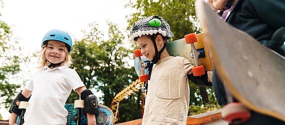 Drei Kinder mit Helmen und Skateboards stehen in einem Skatepark.