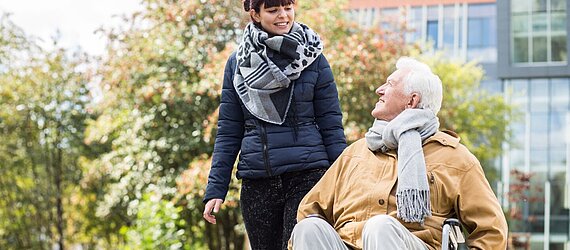 Eine Mitarbeiterin der AWO Mönchengladbach, die hauswirtschaftliche Unterstützung leistet, geht mit einem älteren Mann im Rollstuhl spazieren.