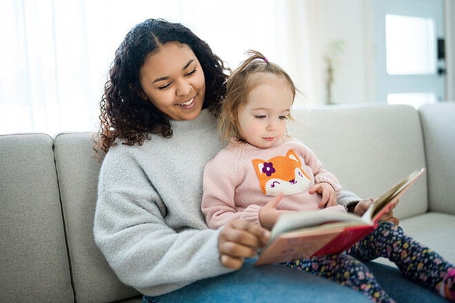 Eine Frau engagiert sich für lebenslanges Lernen, indem sie ihrer Tochter auf der Couch ein Buch vorliest und so Chancengleichheit durch Bildung fördert.